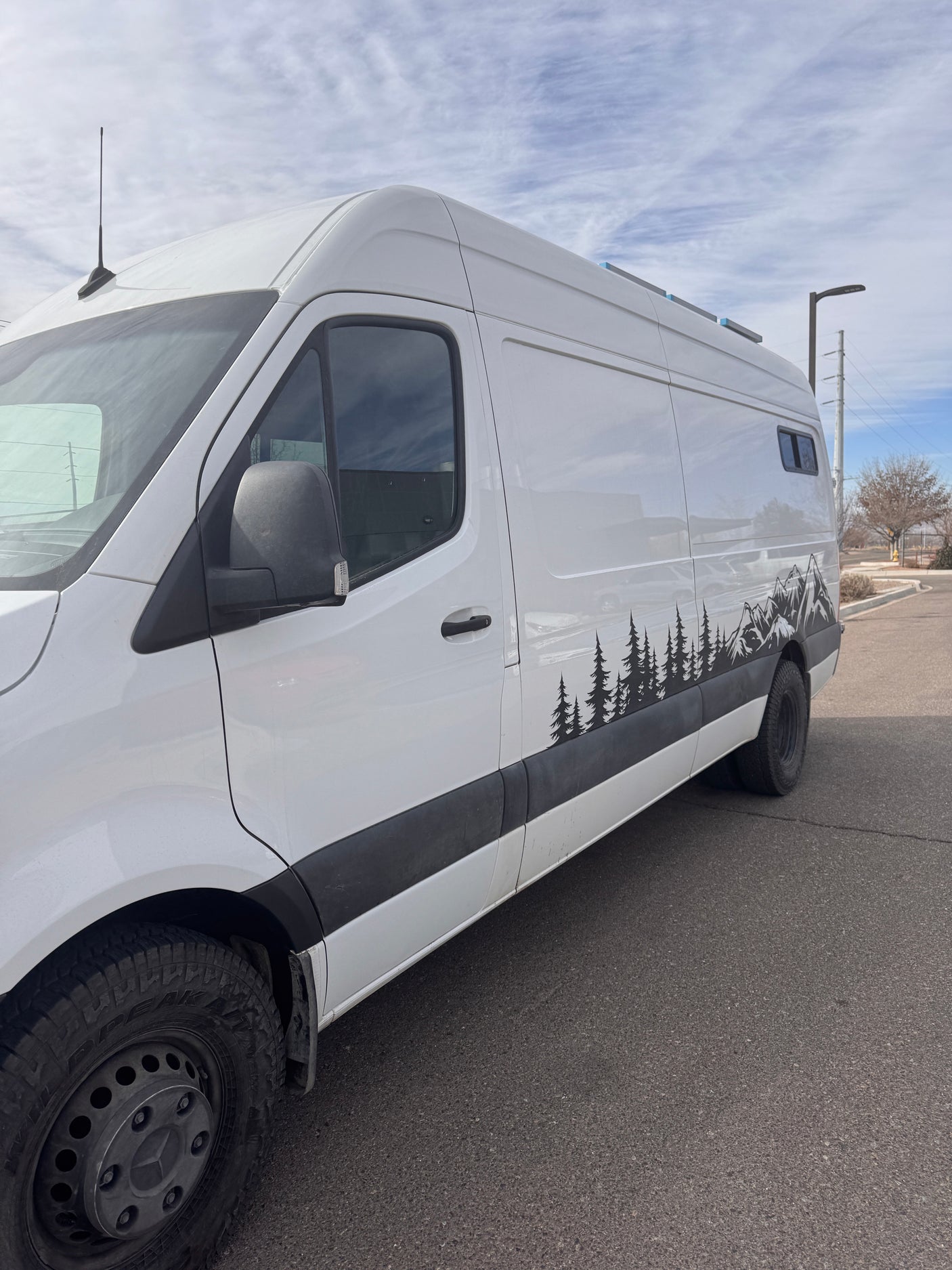 White camper van with nature (trees & mountains) decals on side, parked on asphalt. Ready for your RV rental adventure.