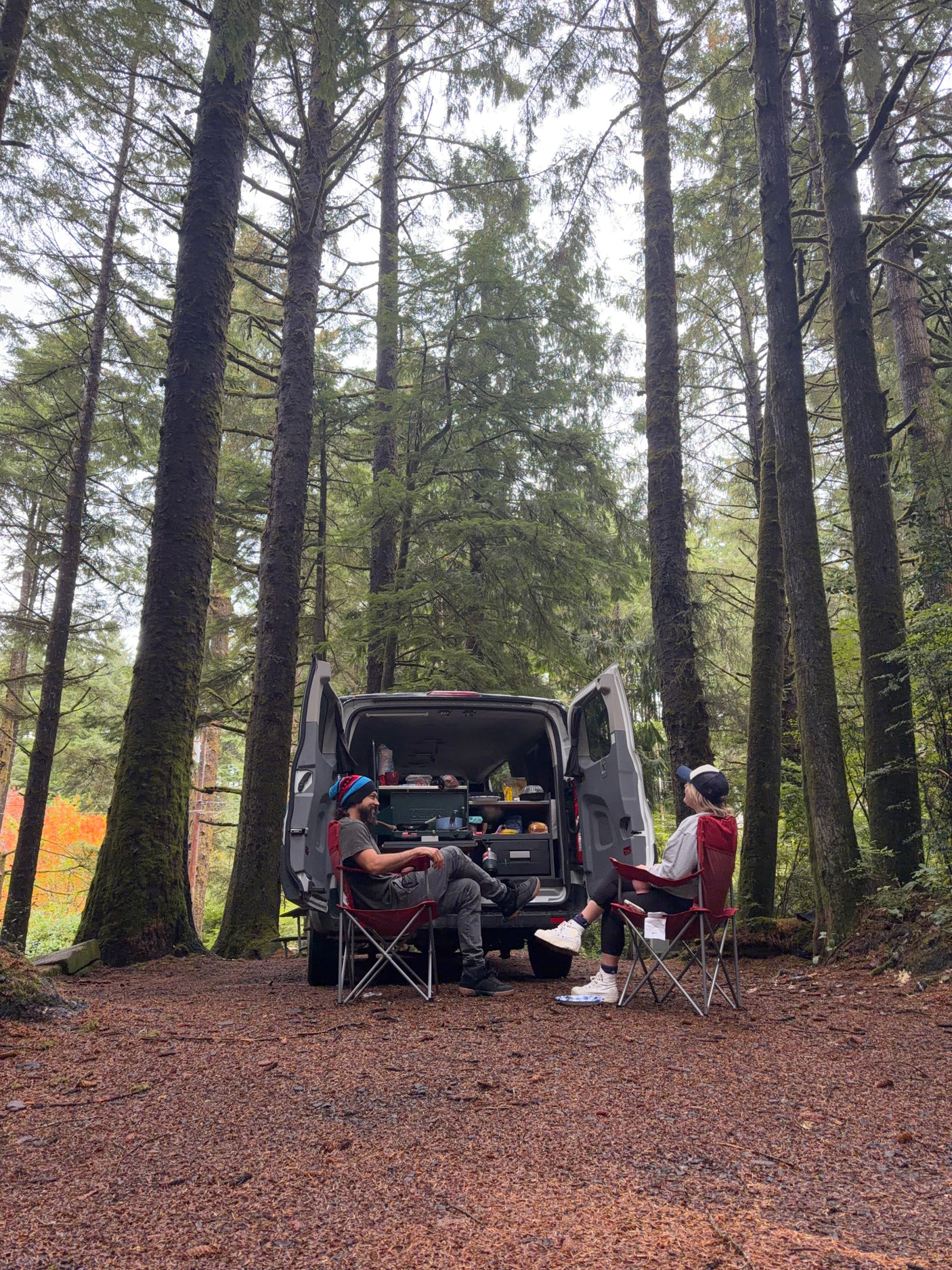 Two people relax by their **rv rental** van, ready for camping in a serene forest with tall trees. Perfect for outdoors.