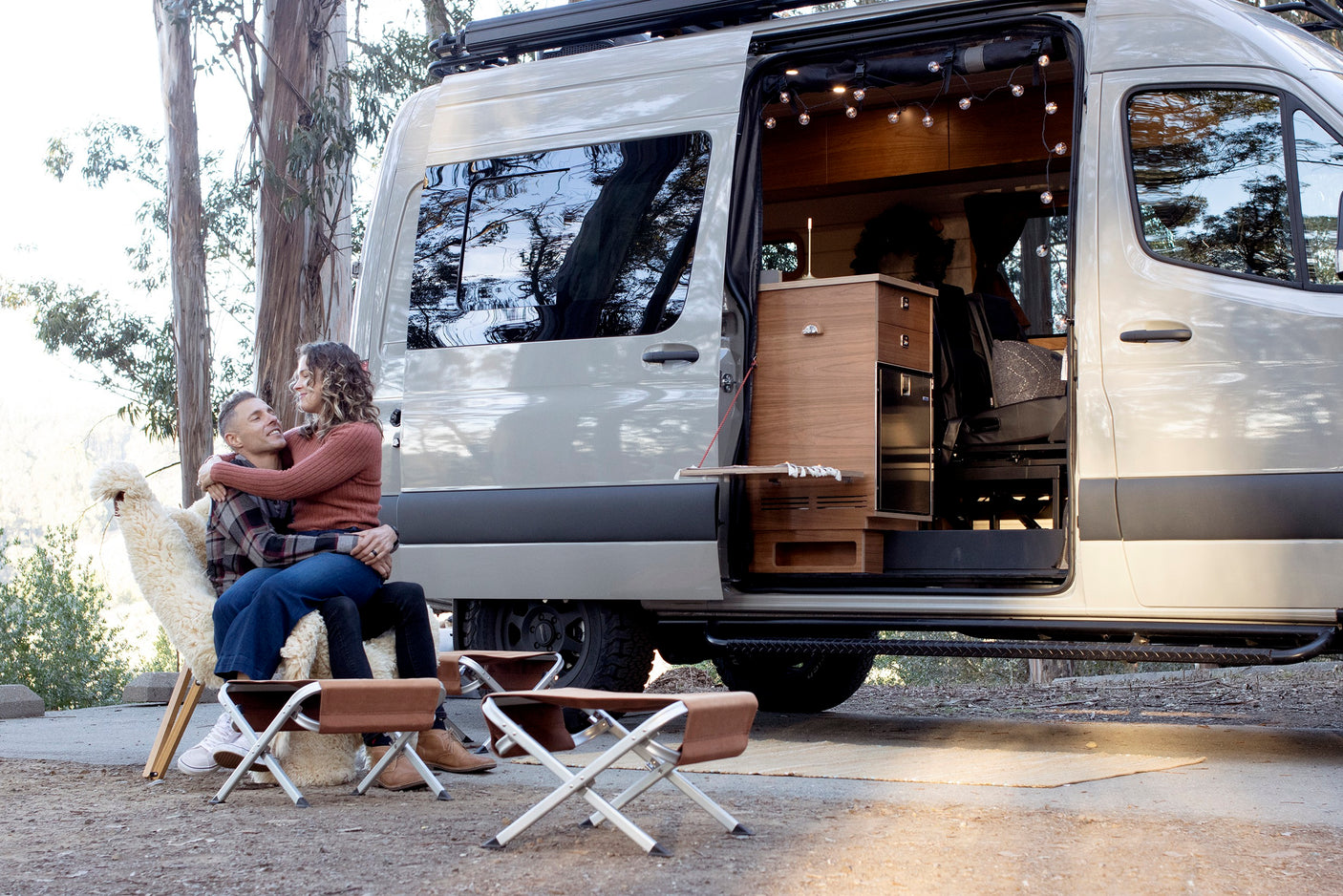 A joyful couple relaxes outside their modern RV rental van with its side door open, revealing a cozy interior.