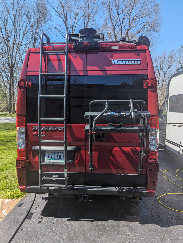 with a blue mat spread on the ground.|Rainy campground scene: a red RV rental with an awning provides shelter. A person is seated at a striped table