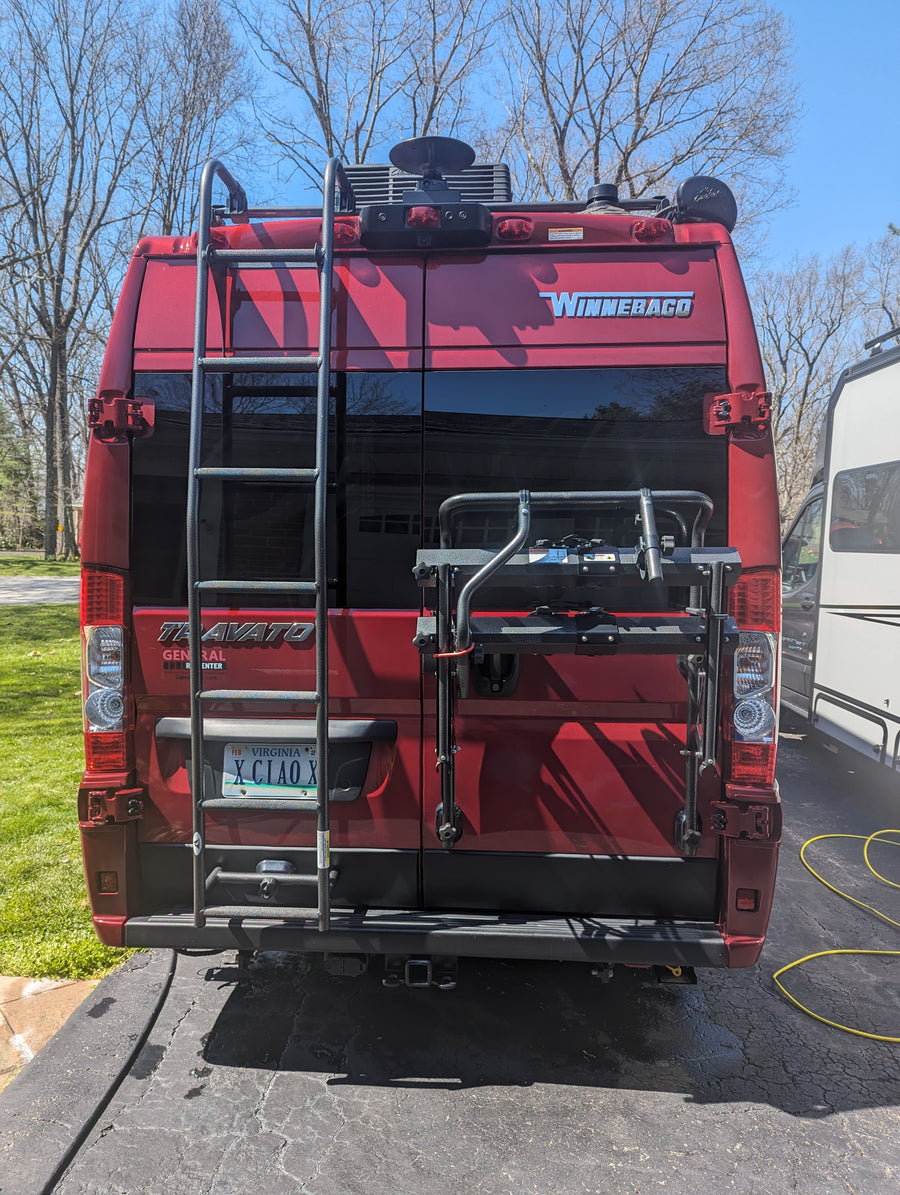 with a blue mat spread on the ground.|Rainy campground scene: a red RV rental with an awning provides shelter. A person is seated at a striped table