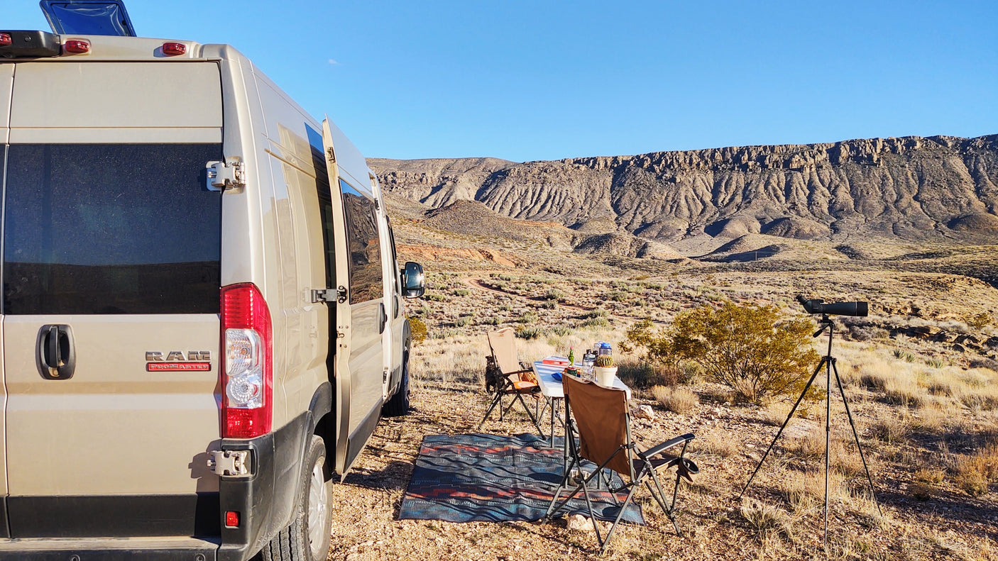Enjoy a scenic RV rental adventure! A tan camper van is parked in a desert landscape with a camp setup & spotting scope.