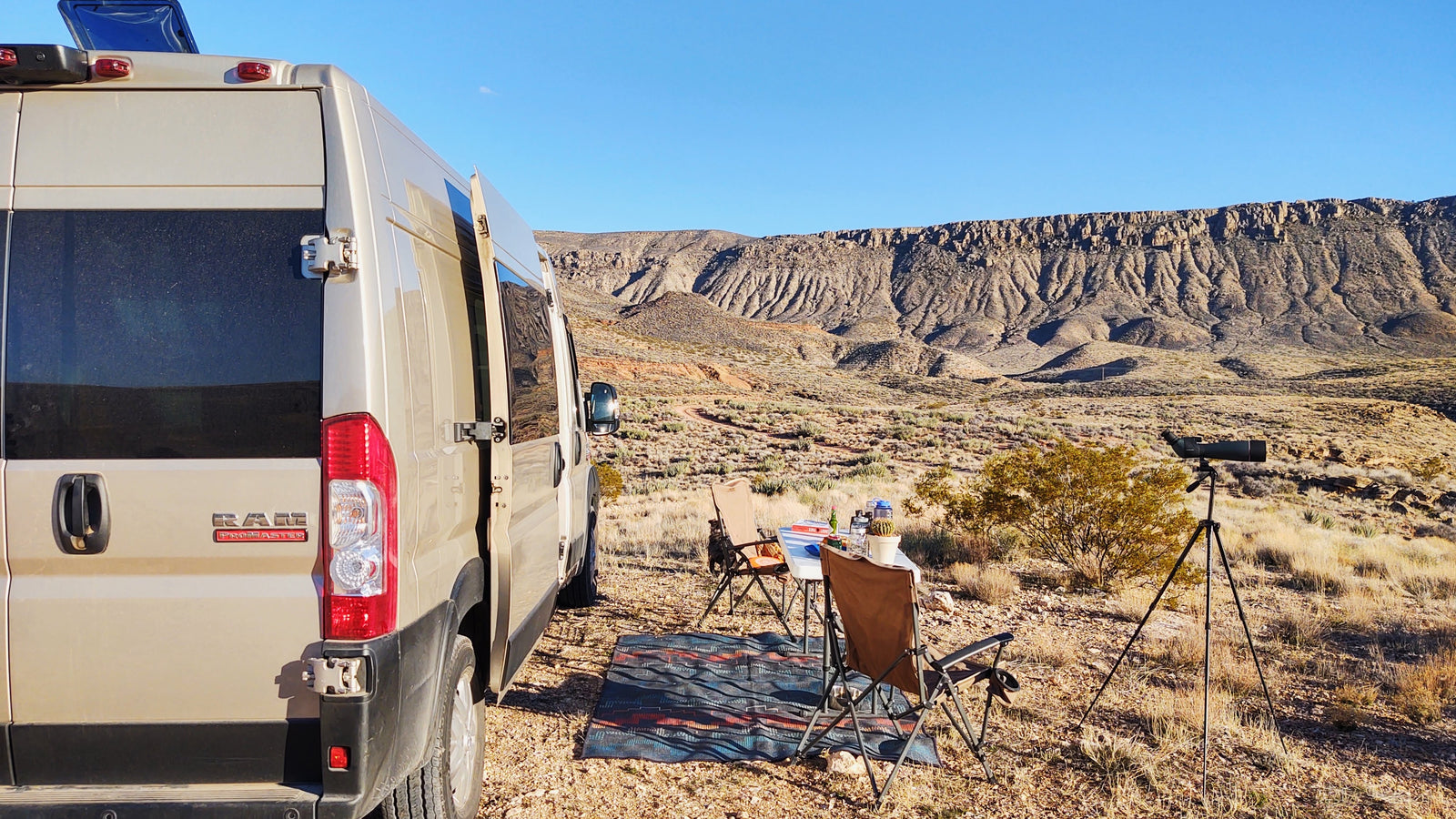 Enjoy a scenic RV rental adventure! A tan camper van is parked in a desert landscape with a camp setup & spotting scope.