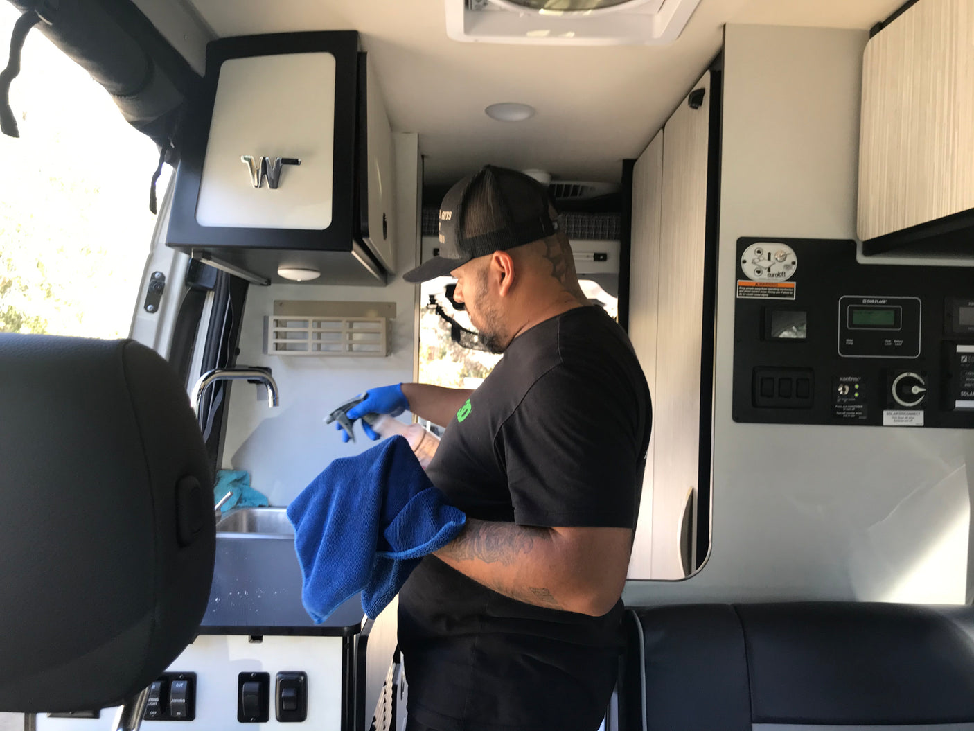 A man in blue gloves cleans an RV sink area with spray and cloth. Typical interior prep for an rv rental camper van.