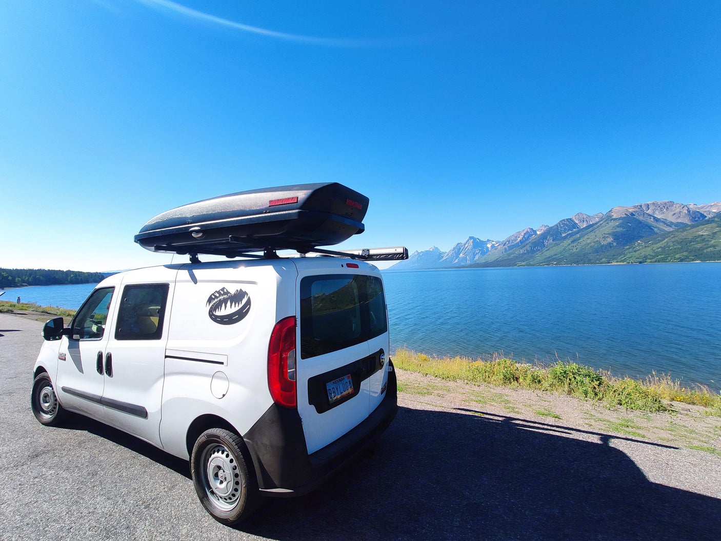 Explore Grand Teton via an RV rental van! White van with cargo box beside a blue lake & majestic mountains.