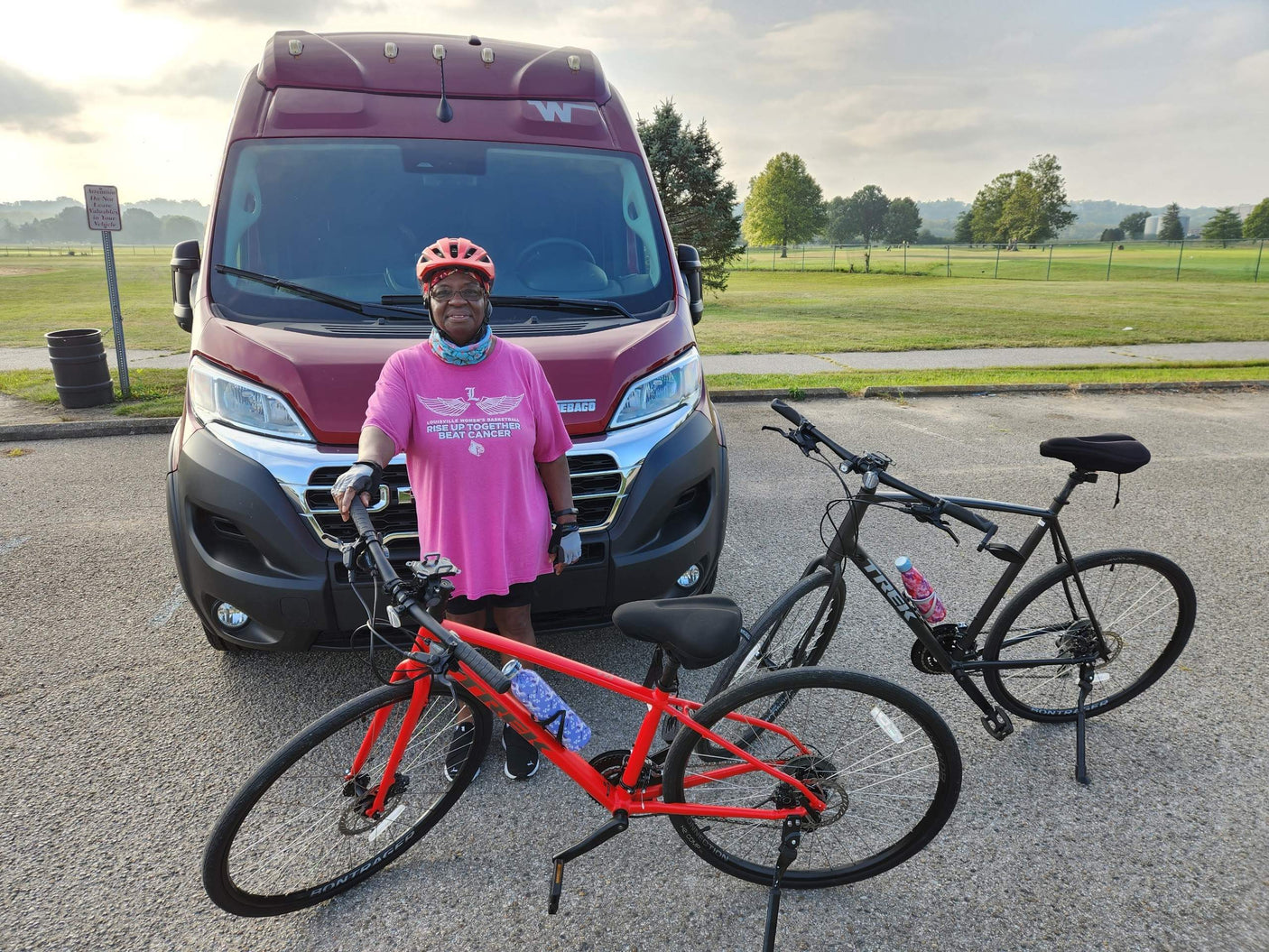 A woman in a pink "Beat Cancer" shirt, bike helmet, and gloves stands by a red RV rental with two bicycles.