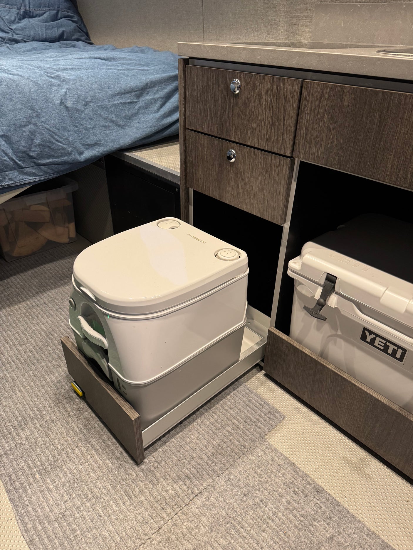 Compact RV rental setup features a Dometic portable toilet on a slide, Yeti cooler, and bed for functional living.