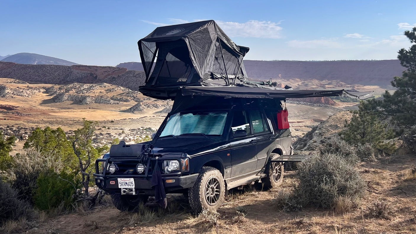Black Land Rover SUV with rooftop tent & awning set up on a dirt path in a scenic desert landscape, like an rv rental.