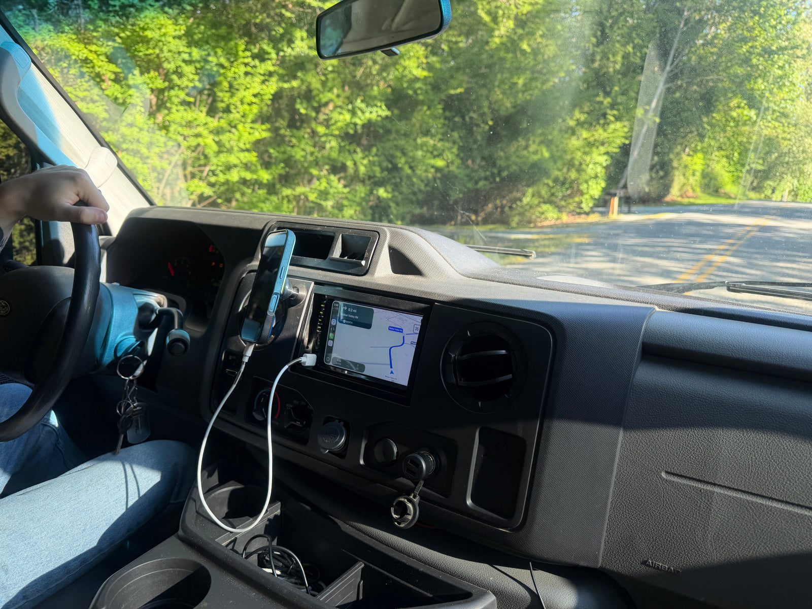 RV rental drive: Navigation on dashboard screen and phone mount. Driver's view of green trees and road ahead.