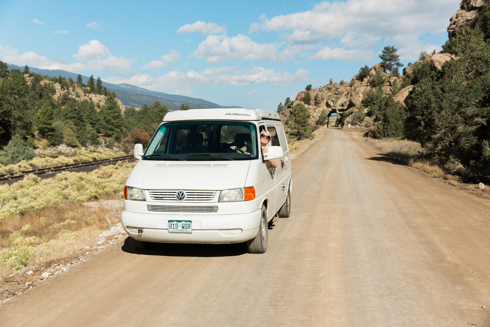 White VW GoWesty RV rental on a dirt road, smiling driver. Mountains, train tracks, and a natural rock tunnel.
