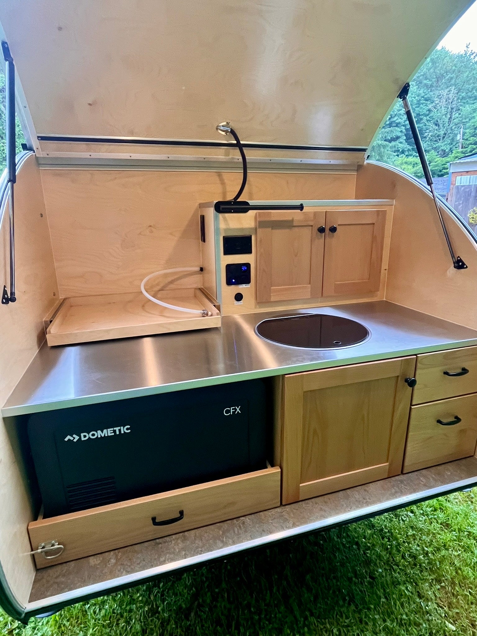 overlooking a green forest and picnic table.|This teardrop RV rental features a red Everest camp stove on a pull-out shelf