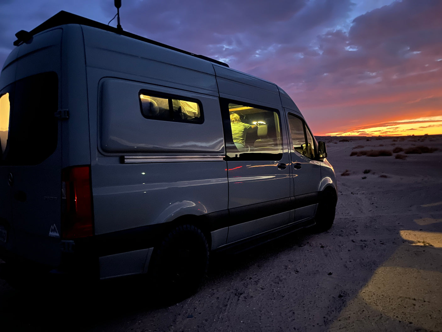 RV rental van parked at stunning desert sunset, interior lit, a person visible inside. Epic van life adventure.