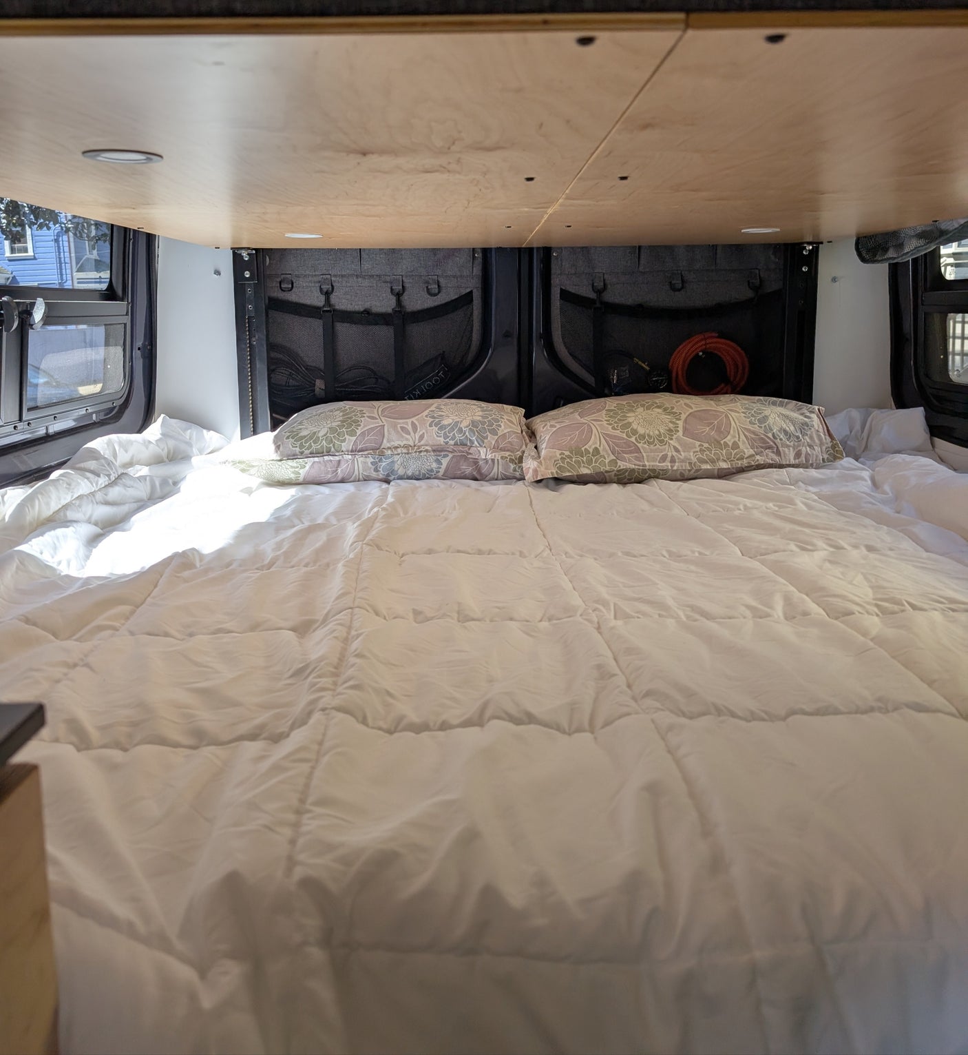 Comfortable RV rental bed with white comforter, patterned pillows, and overhead lighting in a camper van.