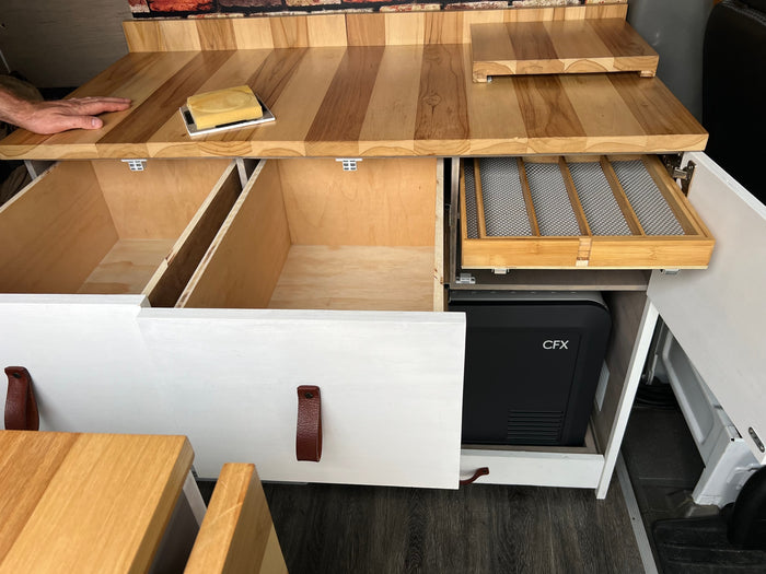 Custom-built RV rental kitchenette with warm wooden countertops, open storage drawers, a cooler, and a clever utensil organizer.