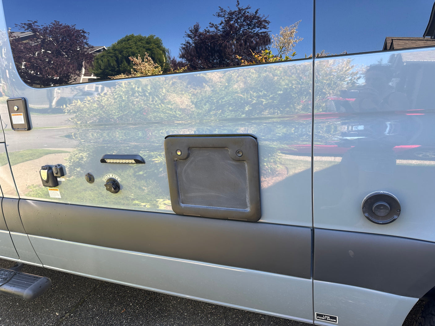 Side of a light gray RV rental showing multiple exterior utility connections, including water fill, electrical, and Truma hot water port.