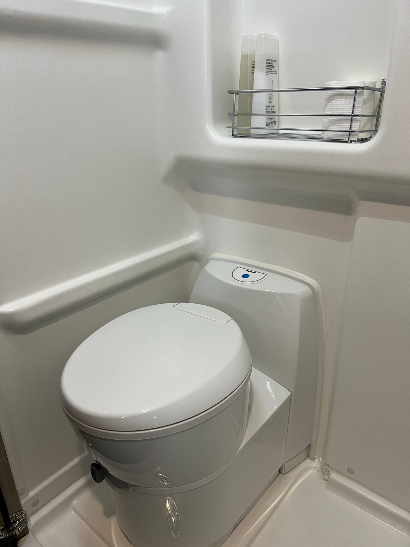 Inside an RV rental: a pristine white bathroom with a modern toilet, shower, and a metal shelf holding essentials.