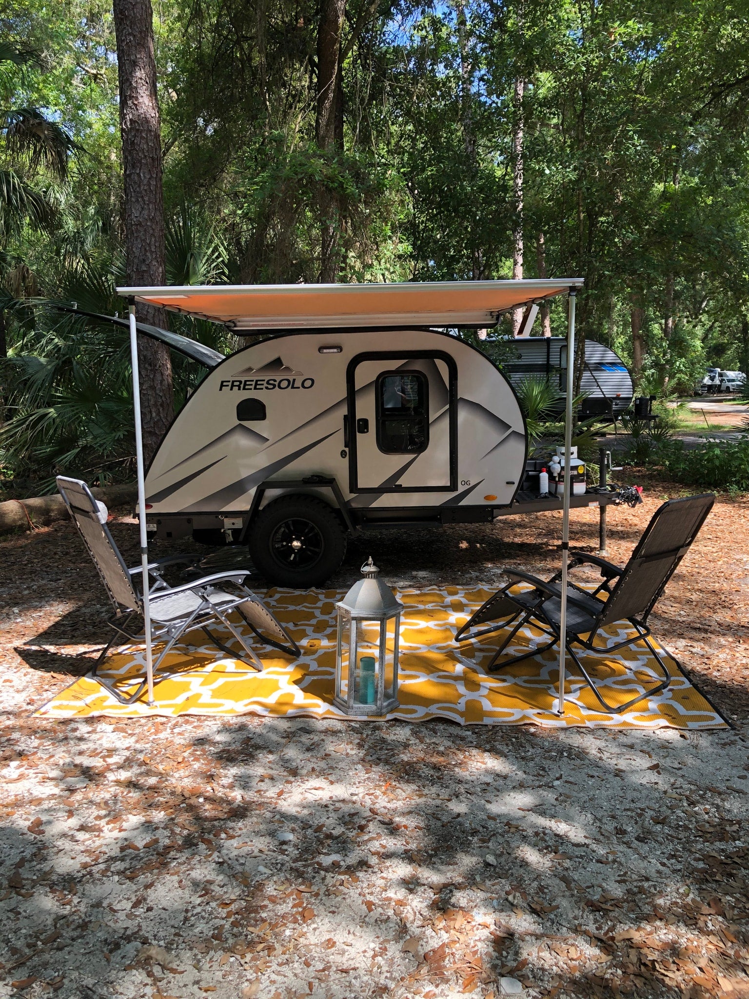 A "FREESOLO" rv rental, a small camper with an awning, set up at a shaded campsite with chairs, a lantern, and a yellow outdoor mat.
