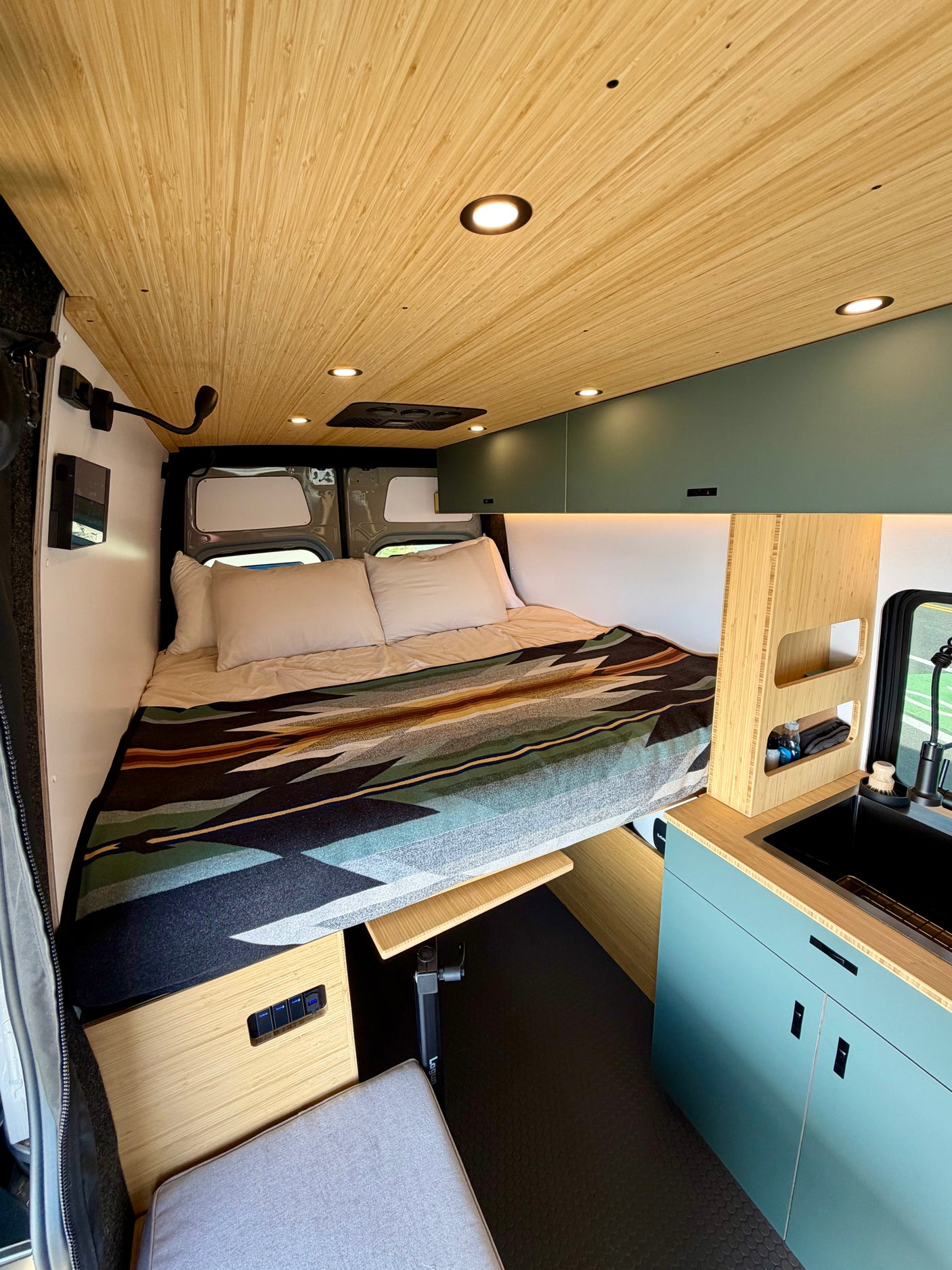 RV rental's modern interior: bamboo ceiling, cozy bed with patterned blanket, kitchenette & smart storage.