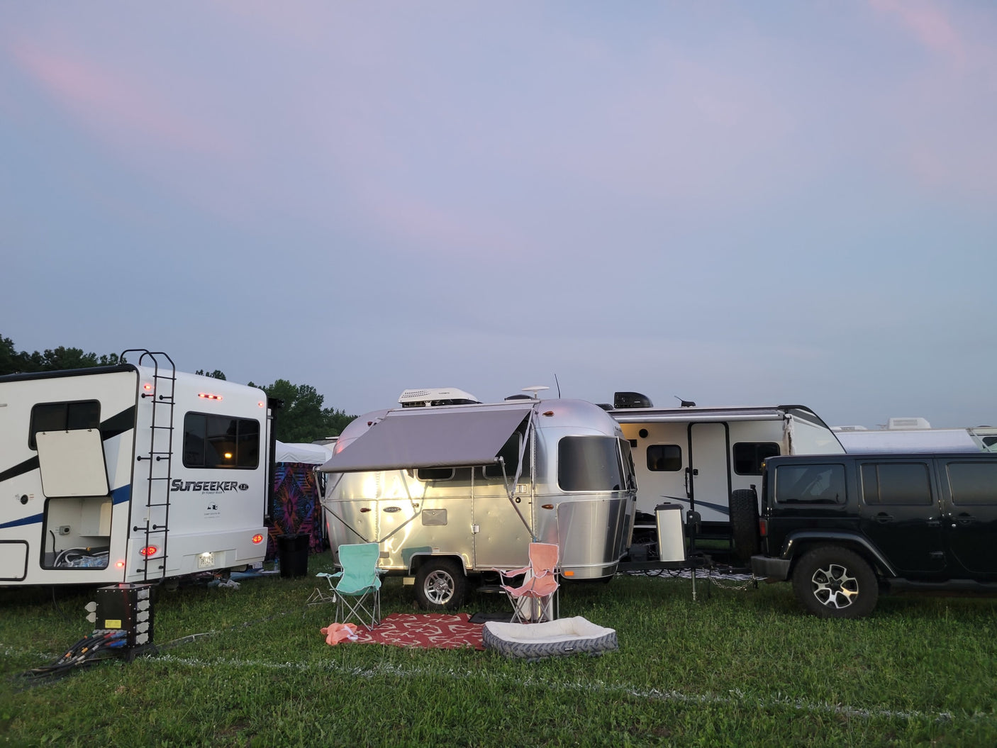RV rental site at dusk: a Sunseeker motorhome, silver Airstream trailer, and Jeep parked on a grassy field.