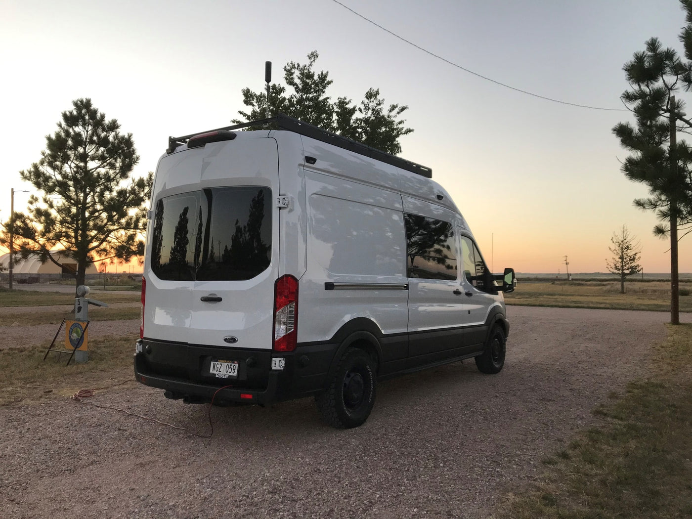 This white camper van, an RV rental, is parked on gravel at sunset, with tall trees and a campsite marker visible. — photo 1