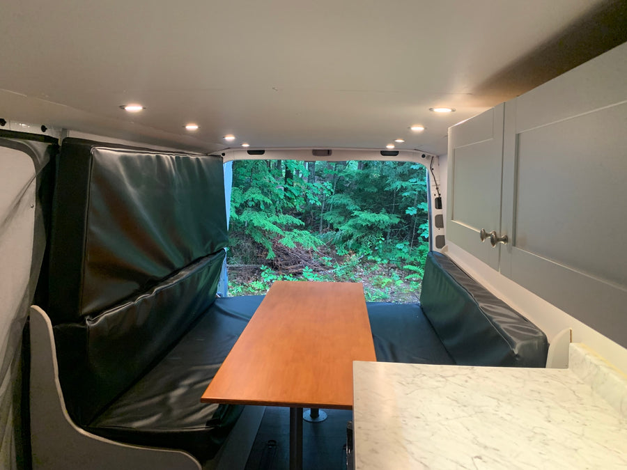 Explore this RV rental's versatile van interior. Features padded benches, dining table, storage, and a forest view.