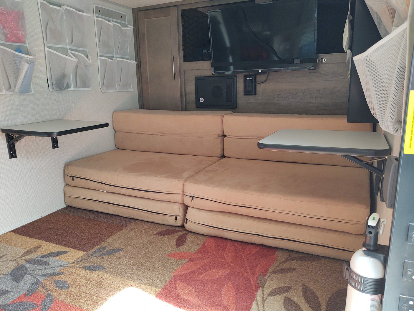 Interior of an RV rental featuring modular seating, two fold-down tables, TV, mesh storage, and a fire extinguisher.