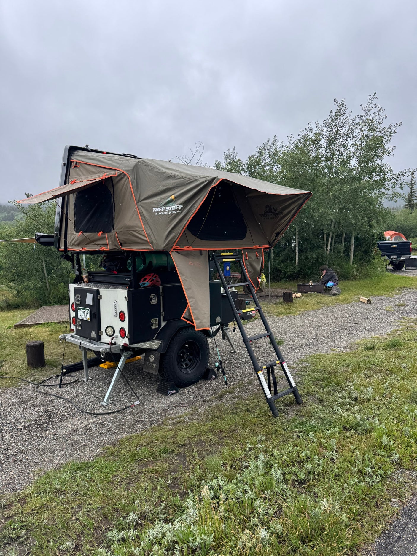 Experience overland camping with this RV rental! A gray trailer with olive rooftop tent and ladder, set up at a gravel site.