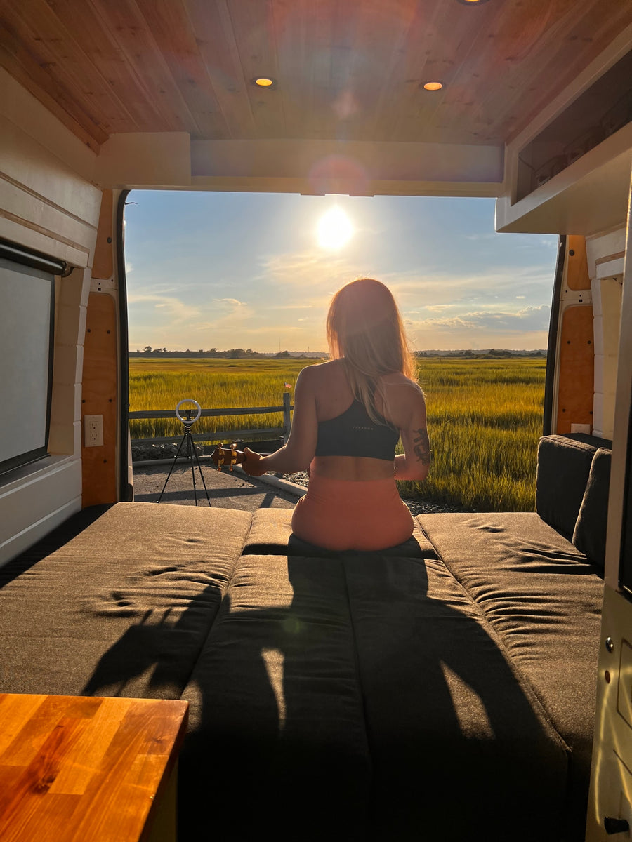 Inside an rv rental, a woman sits on a bed, playing ukulele, facing a stunning sunset over golden fields.