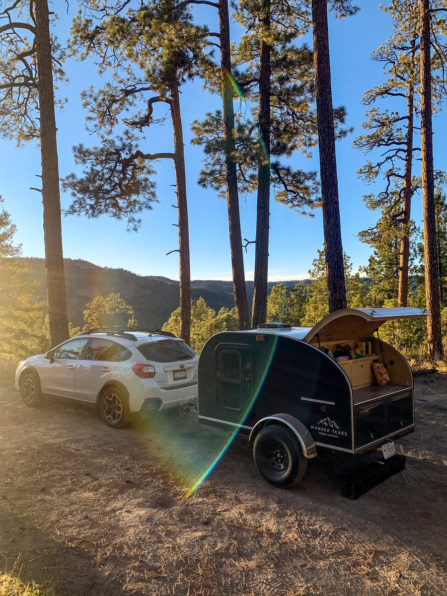 White Subaru Crosstrek and a black Wander Tears teardrop RV rental at a sunny forest campsite