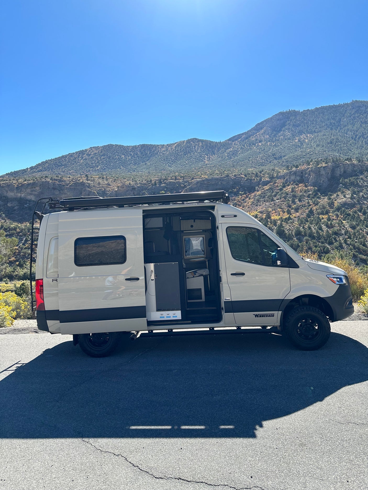 RV rental camper van parked on a mountain road with its side door open, revealing the interior under a clear blue sky.