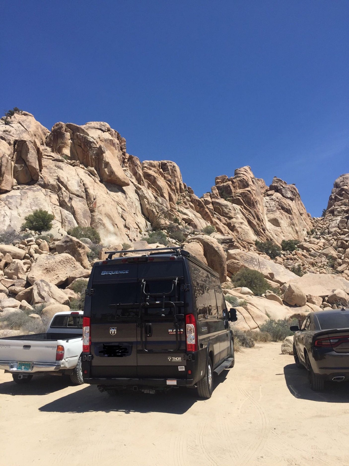 Exploring rocky terrain: An RV rental van, truck & car parked amidst large desert boulders under a clear sky.