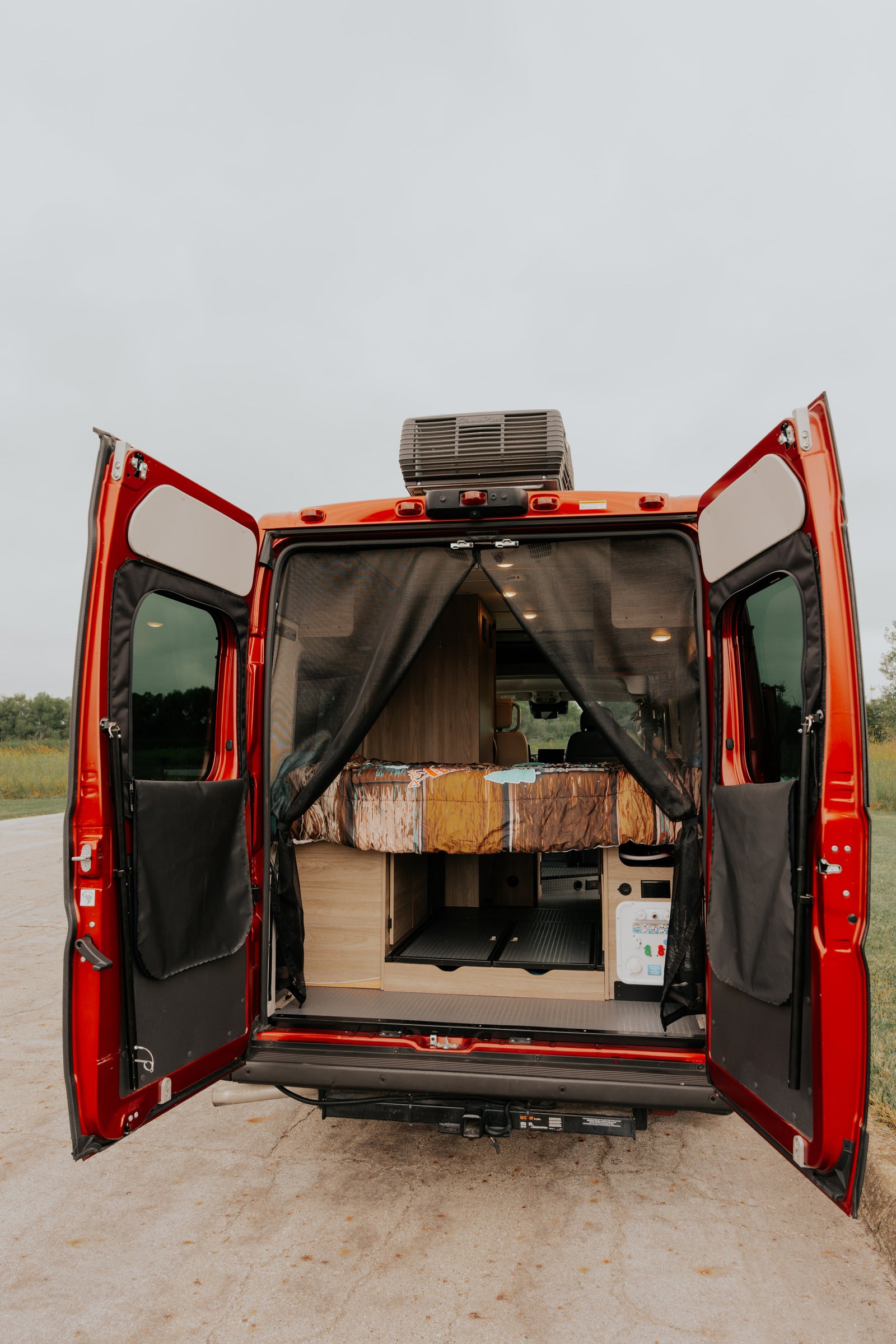 Red camper van for rv rental, rear doors open revealing bed, storage, and water hookups inside.