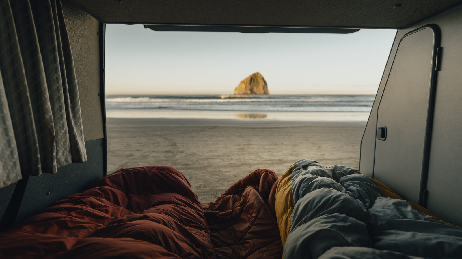 Inside an RV rental, two sleeping bags face a serene beach with a large sea stack in the ocean.