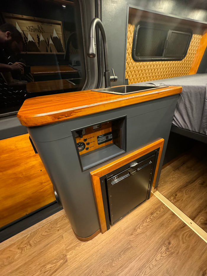 Explore this RV rental's cozy interior: a sleek kitchen area with sink, fridge, and wooden counter, next to a comfy bed.