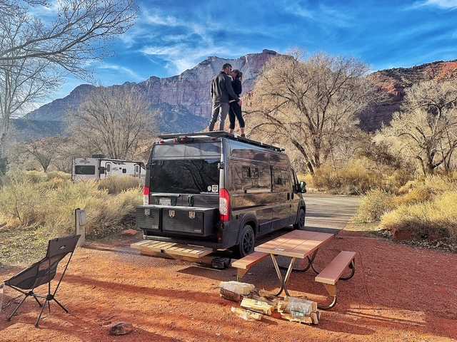 enjoying ocean views from his solar-powered setup.|Couple kisses atop their RV rental at a scenic mountain campsite with a picnic table & firewood. Perfect outdoor adventure!|Dark gray camper van with rooftop solar & bike rack