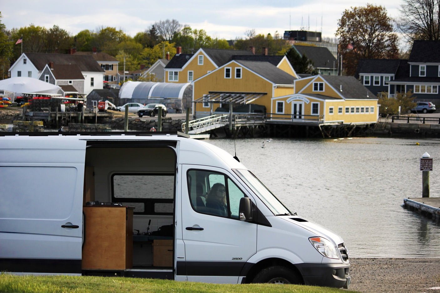 White RV rental van at waterfront; a person is inside its cozy camper conversion, ready for adventure.