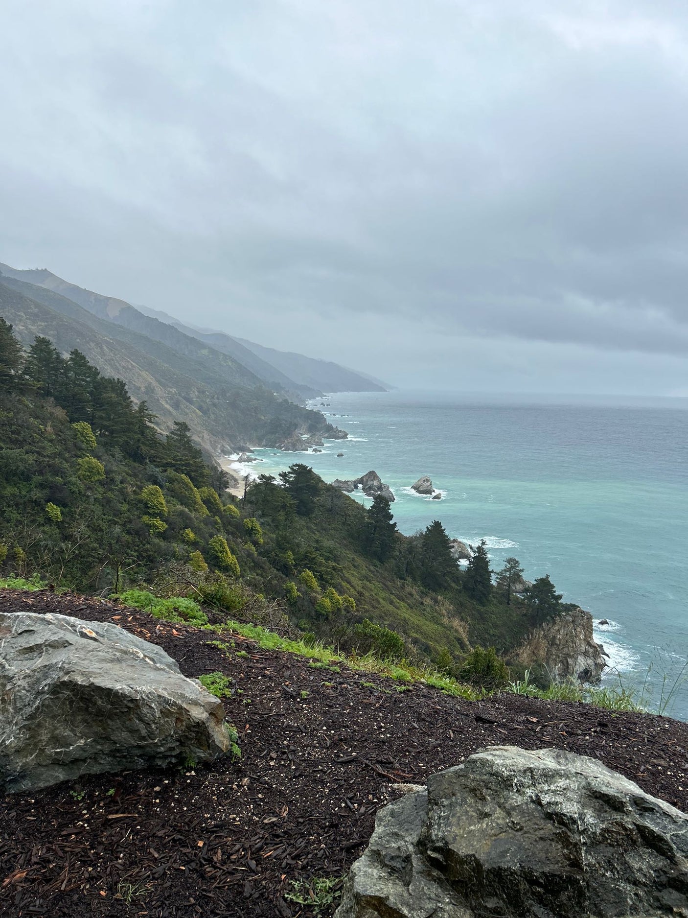 RV rental ideal for Big Sur's dramatic coast: A misty view of lush green mountains meeting turquoise ocean under an overcast sky.
