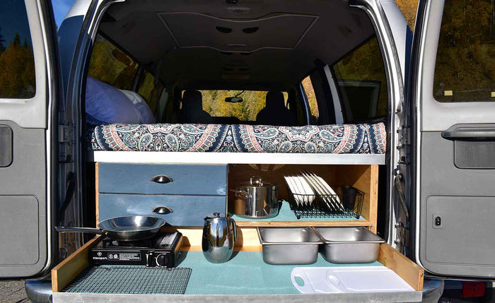 This rv rental features a cozy bed above a pull-out kitchen with stove, pan, and dishes, perfect for camper adventures.