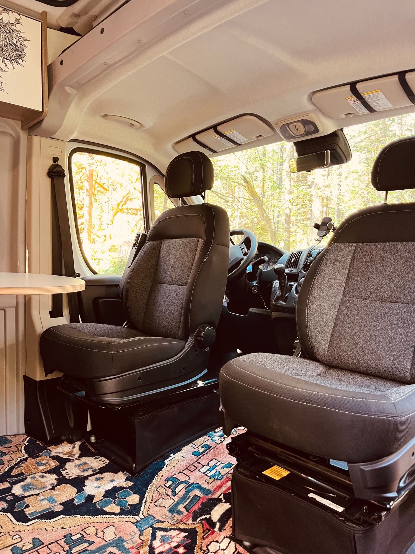 Modern RV rental interior with swivel seats, a small table, and a vibrant rug. Trees visible through windows.