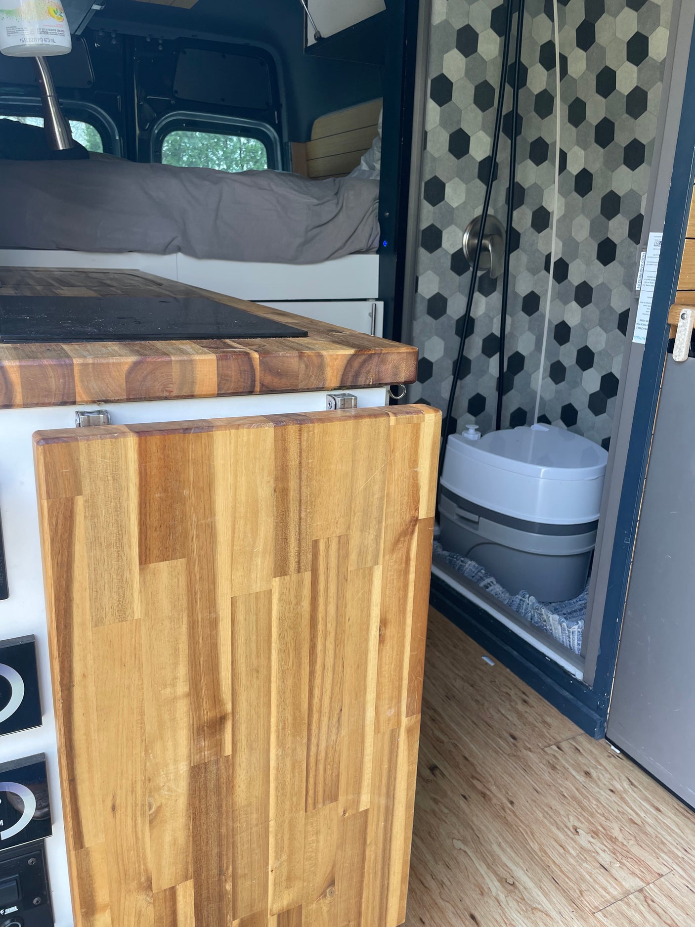 Compact RV rental interior featuring a bed, kitchen counter with cooktop, and a convenient portable toilet in the shower.