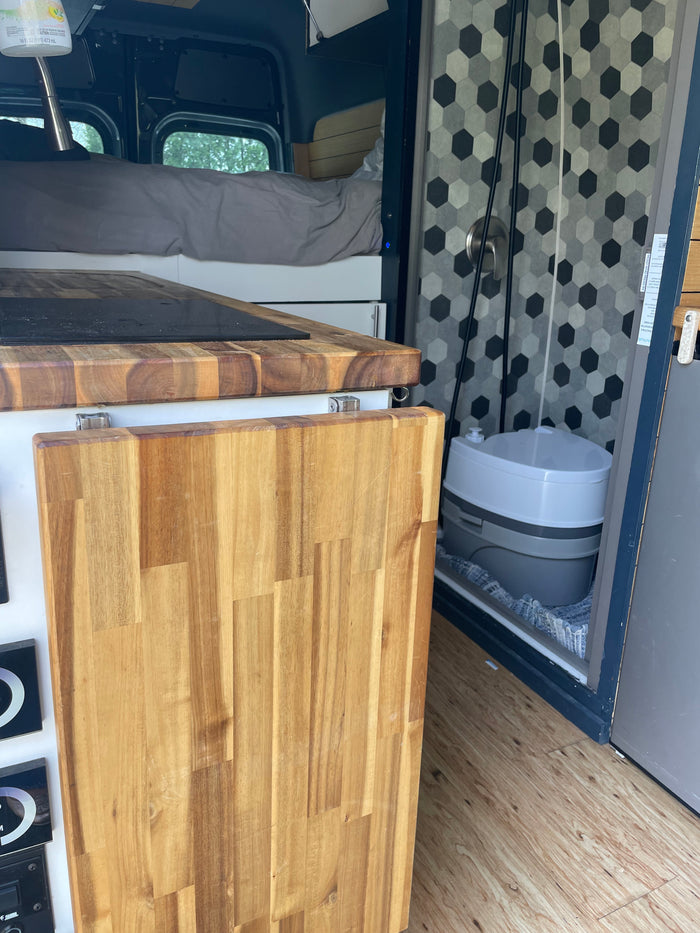 Compact RV rental interior featuring a bed, kitchen counter with cooktop, and a convenient portable toilet in the shower.