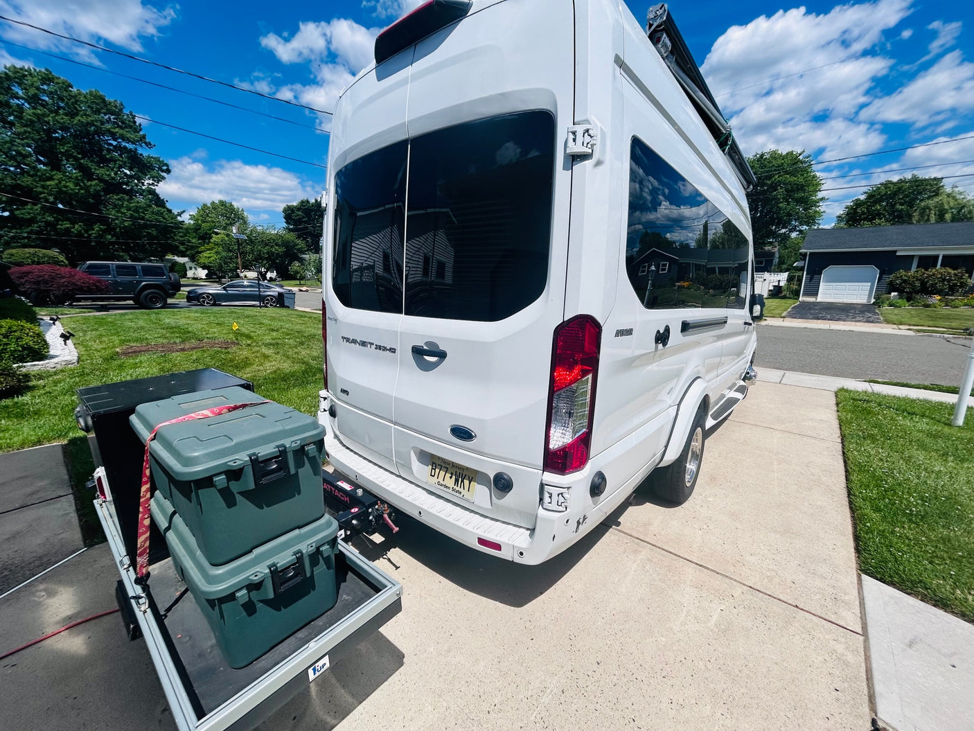 White Ford Transit RV rental parked on a suburban driveway, with gear secured on a rear cargo carrier under a sunny sky.