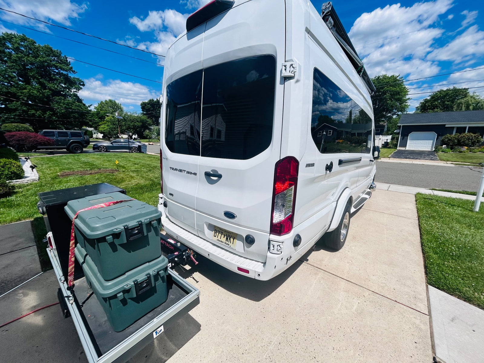 White Ford Transit RV rental parked on a suburban driveway, with gear secured on a rear cargo carrier under a sunny sky.
