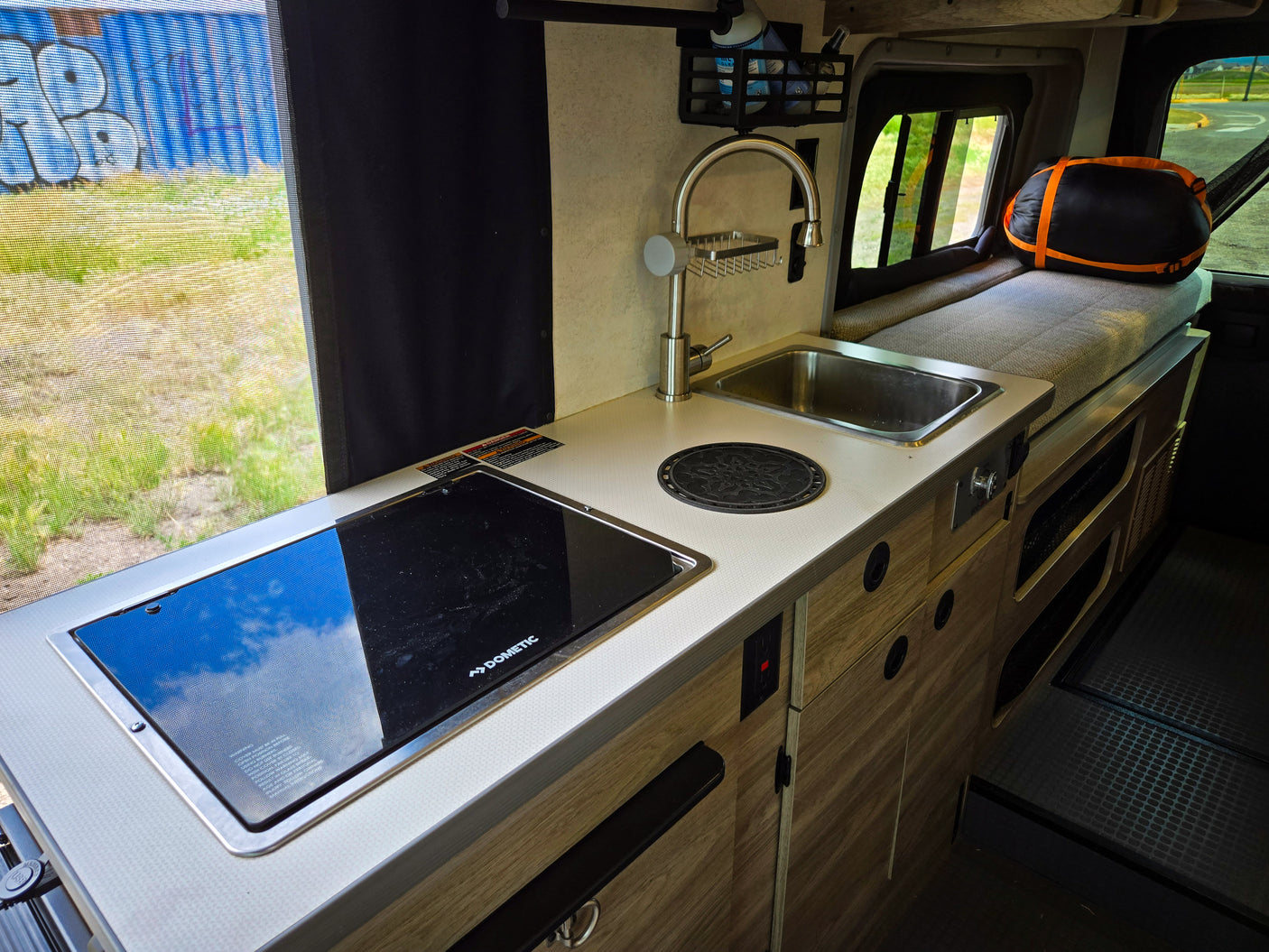 Inside an RV rental: a compact kitchenette with sink, Dometic cooktop, and cozy bench bed. Window views show outdoor scenery.