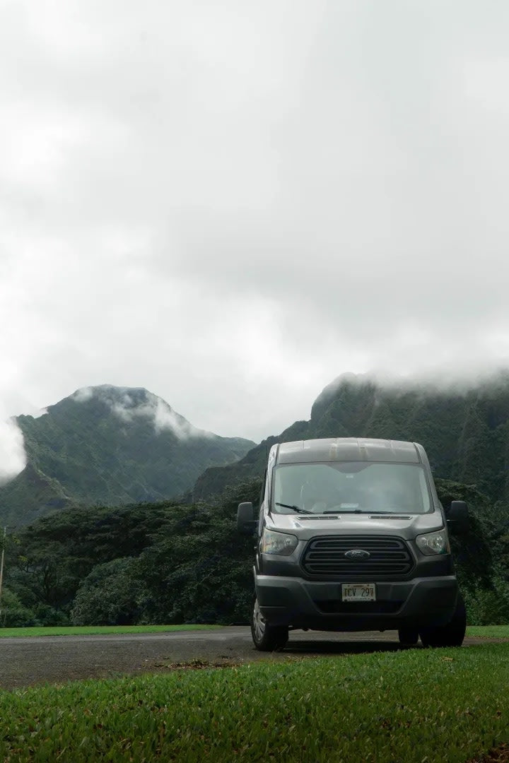 Grey Ford Transit van, an RV rental, parked amidst misty green mountains. Perfect for scenic travels.