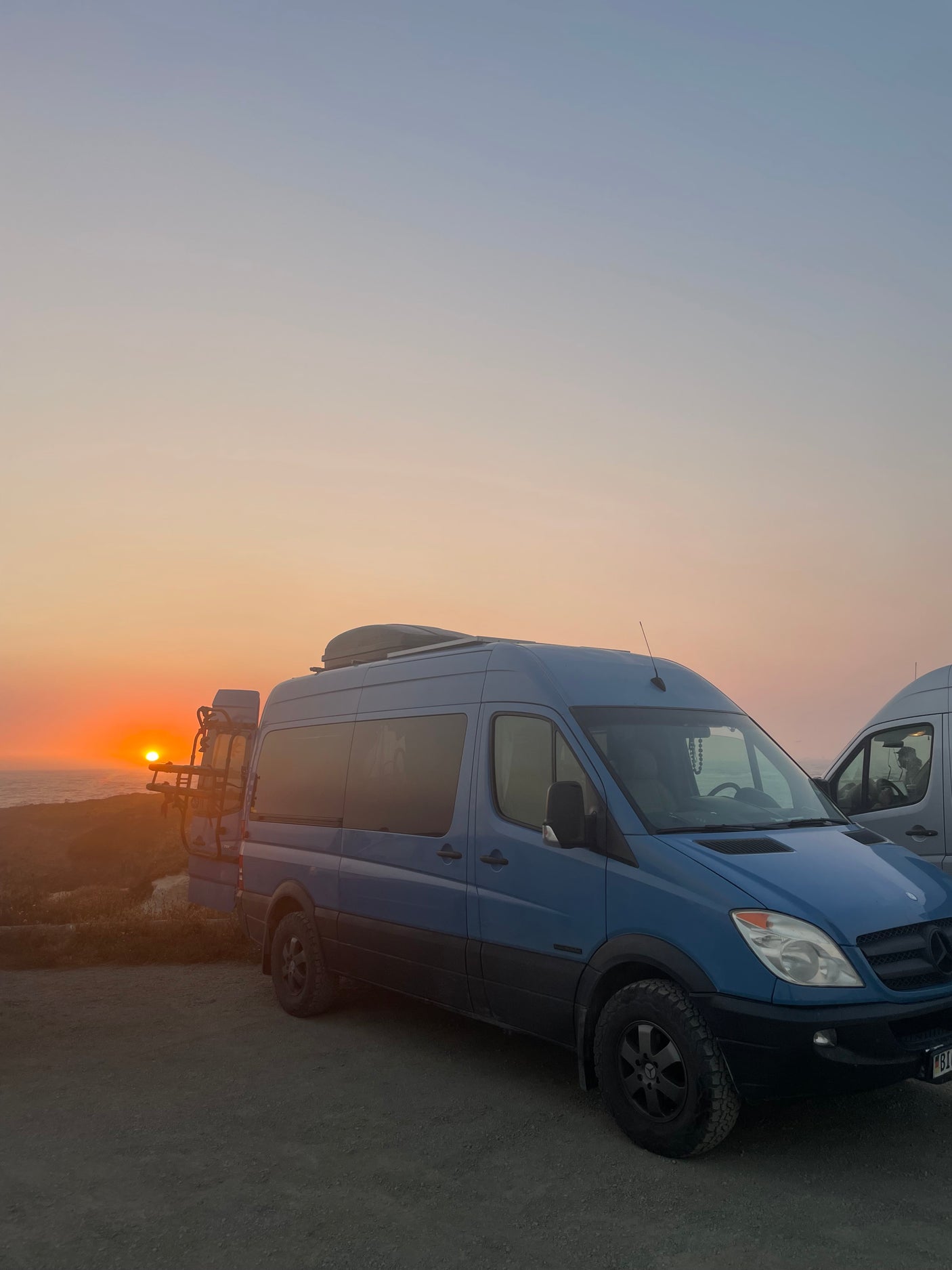 Scenic RV rental: Blue Sprinter van at sunset over the ocean, bikes on rear. Ideal for adventure.