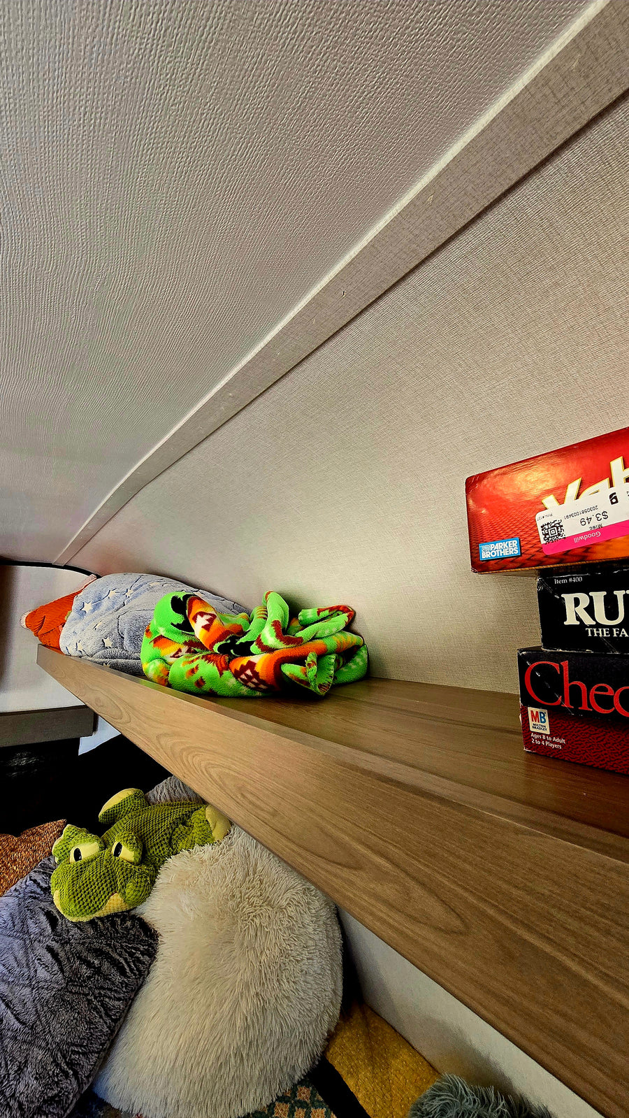 RV rental overhead shelf with games (Goodwill price tag visible), a colorful blanket, and plush toys in cozy storage.