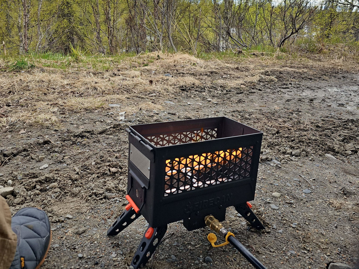 A black portable propane fire pit with orange legs burns brightly on a gravel campsite, perfect for an RV rental outdoor adventure.