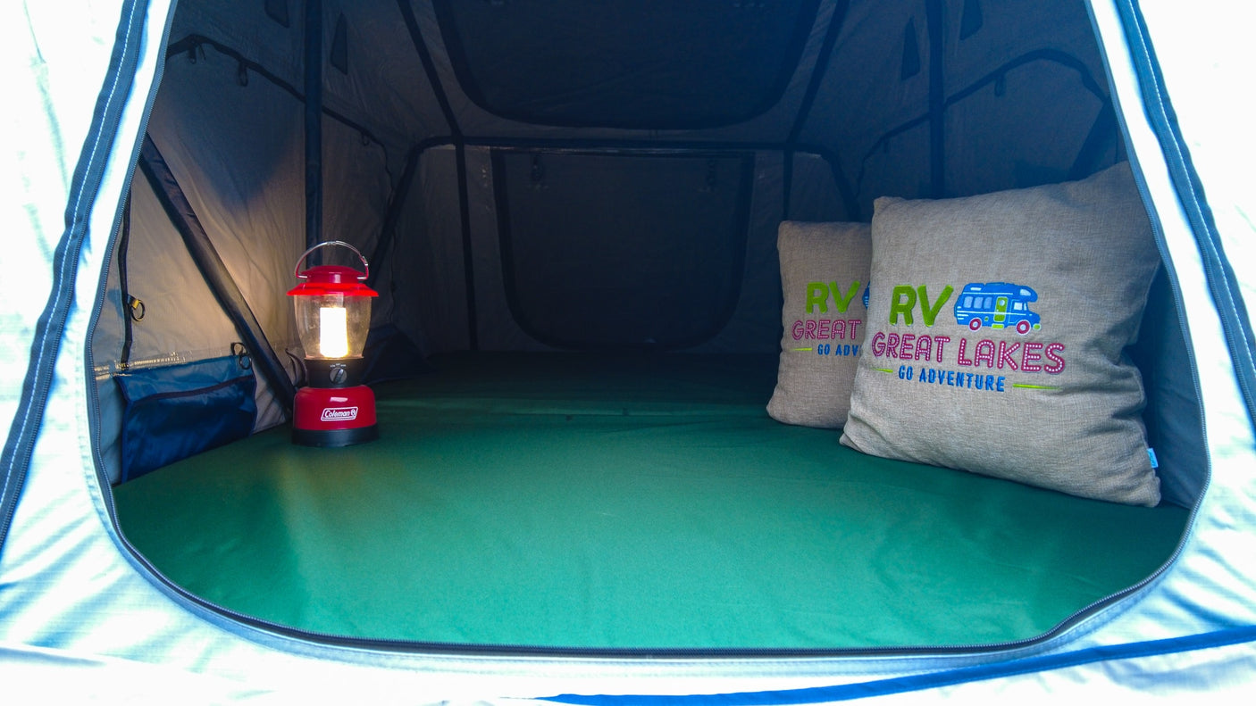 Inside an RV rental tent: a Coleman lantern, comfy green bed, and "RV Great Lakes" pillows await your adventure.