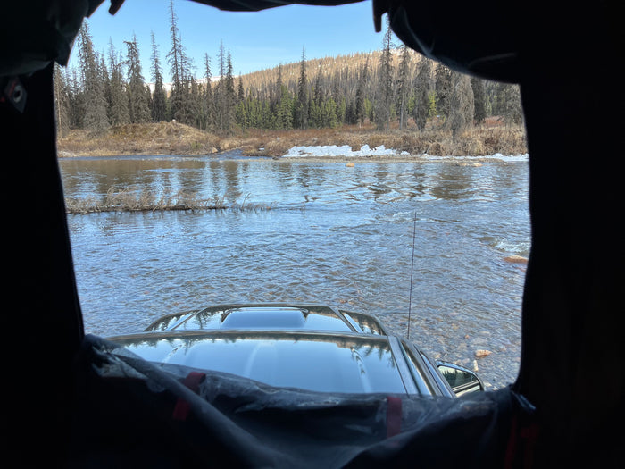 View from an RV rental's window: a vehicle crossing a shallow river, surrounded by a winter forest.